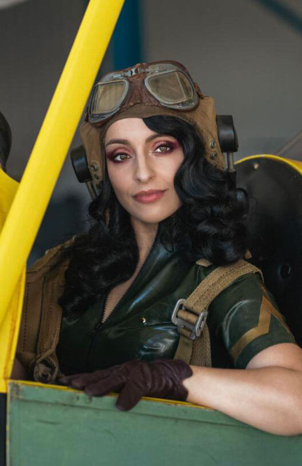 Person in a vintage pilot costume with goggles inside a yellow and green vehicle. Black 1940s style wig with long marcel waves: Vivienne