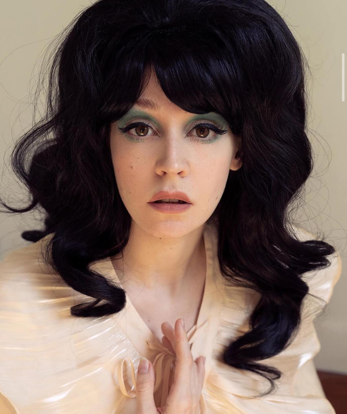 Person with dark wavy hair and dramatic makeup wearing a light-coloured outfit. Long black high crown wig, curly: Brigitte