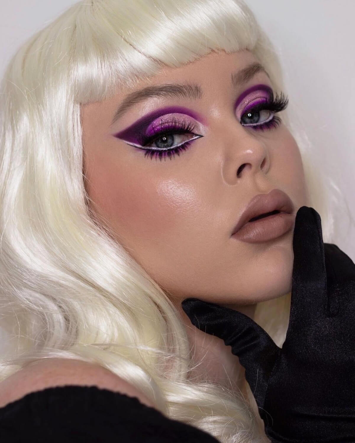 Person with blonde wig and dramatic makeup, wearing a black top. Platinum blonde pinup style wig with short fringe: Eira 