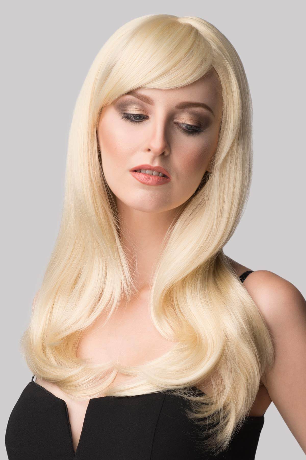 Woman with long blonde wig on a gray background. Long blonde wig, face framing with long, side-sweeping fringe: Ashley blonde 613