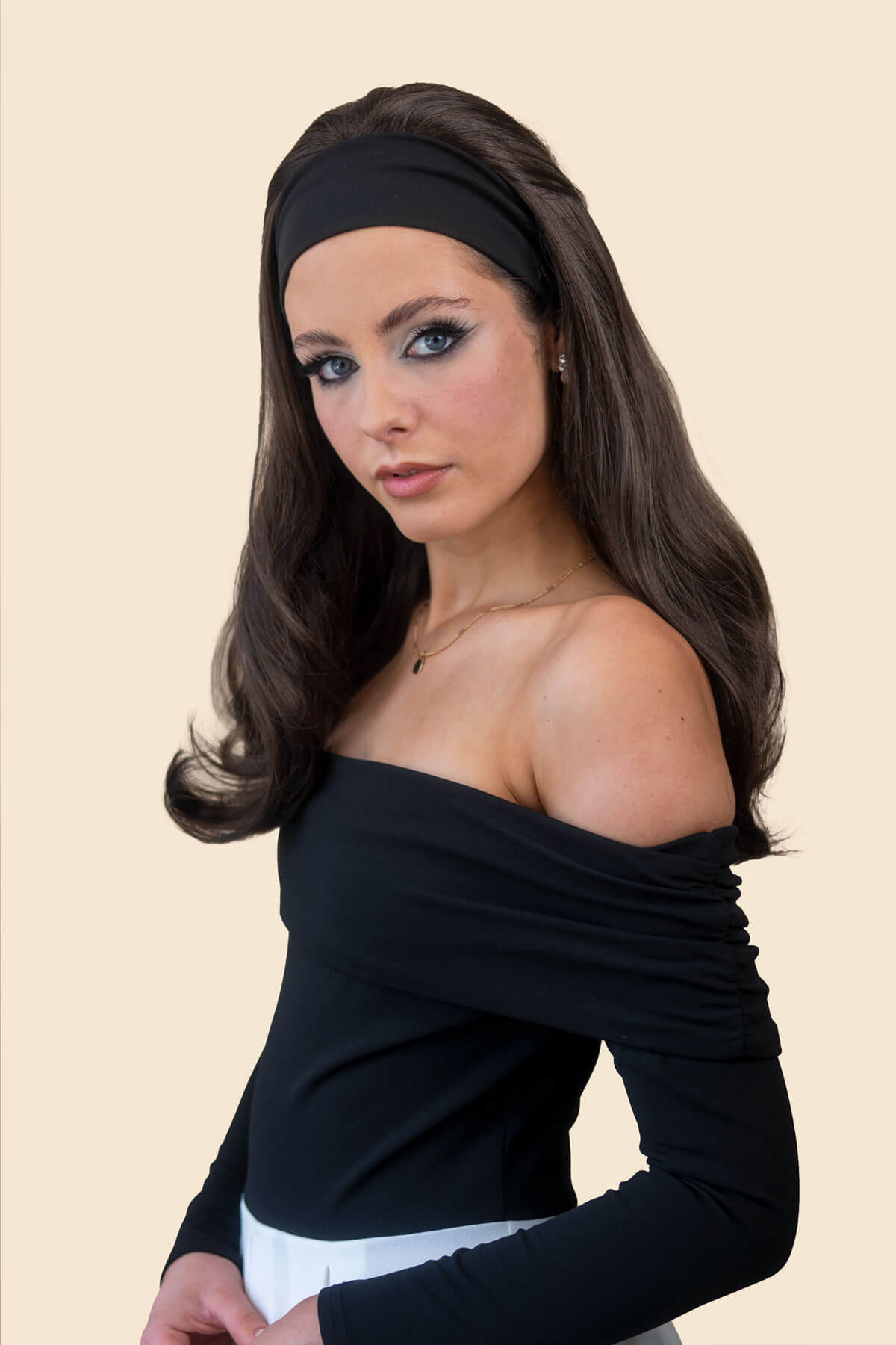 A woman with long dark brown hair wearing a black off-the-shoulder long-sleeve top, a black headband, and a white bottom, posed against a solid light beige background. Long Gently Layered Half Wig with curls towards the tips: Jessica black 1b