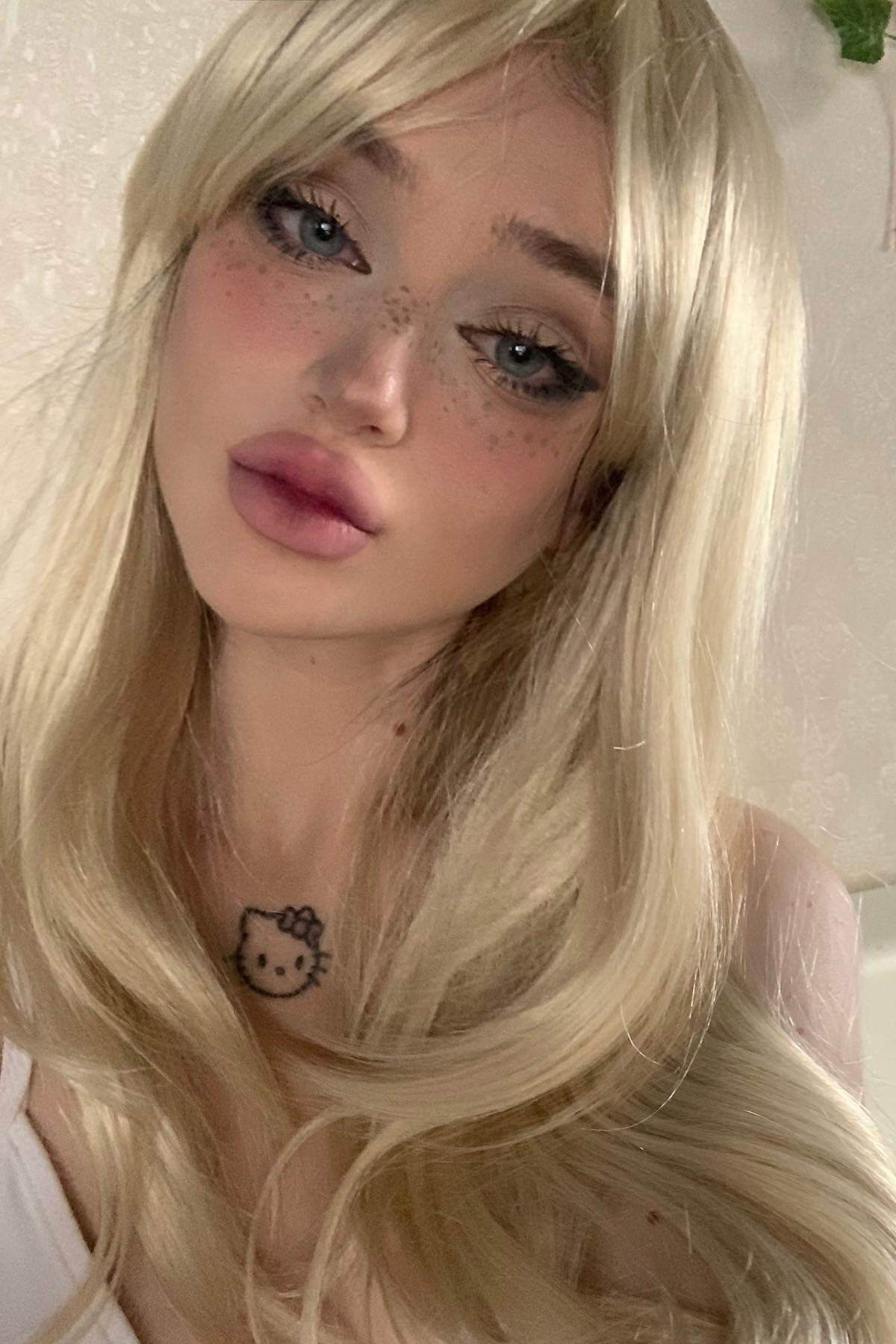 Person with long blonde hair and a Hello Kitty tattoo on their neck, against a neutral background. Long blonde wig, face framing with long, side-sweeping fringe: Ashley blonde 613