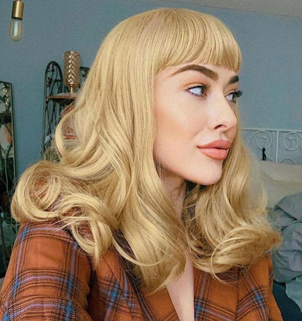 A woman wearing a blonde wavy wig with straight bangs, styled in a retro 1960s or 1970s look, paired with a brown plaid shirt. Ash blonde 50s style wig with short fringe and gentle waves: Trinity