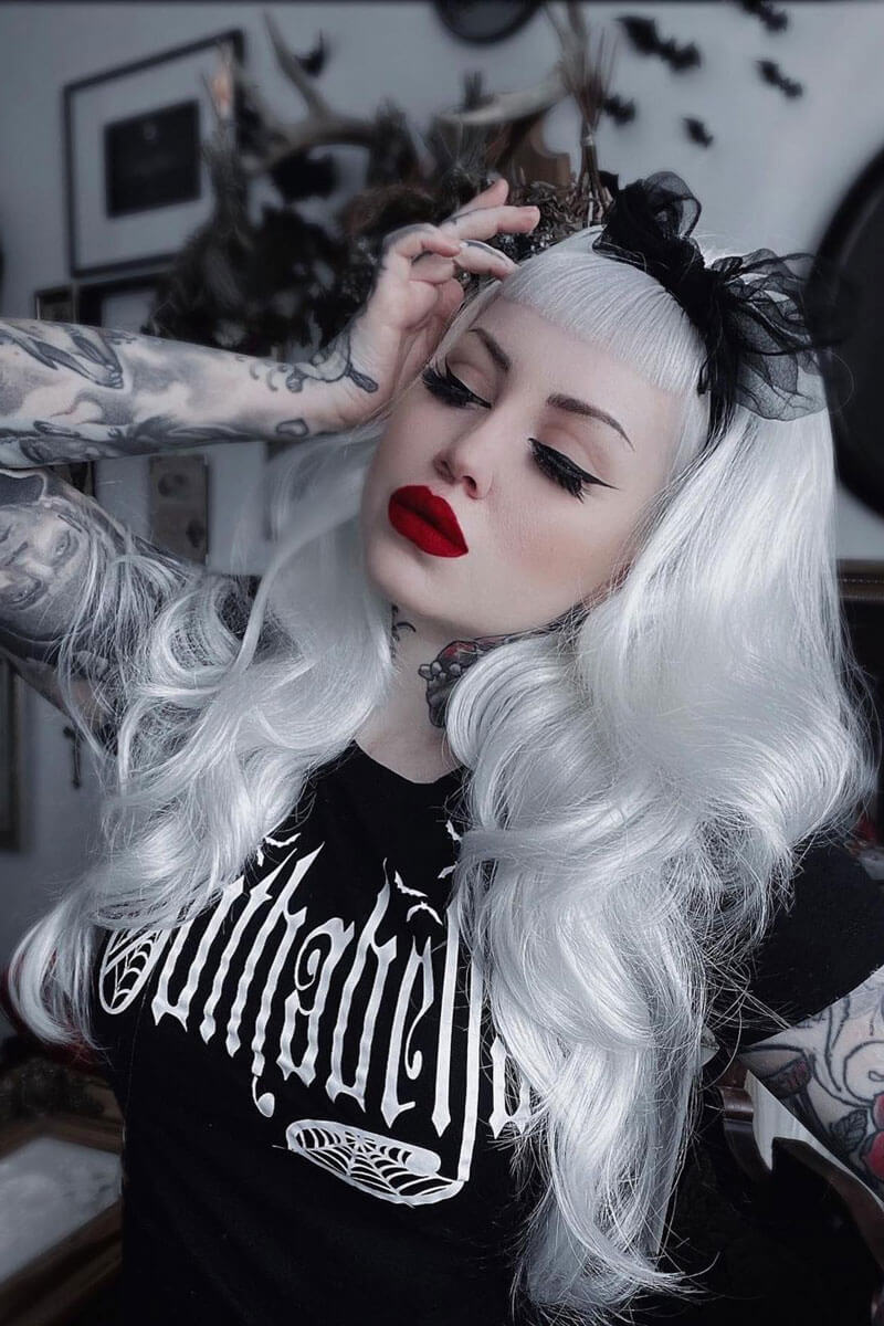 Person with white hair and red lipstick wearing a black 'Vampira' shirt in a darkly decorated room.