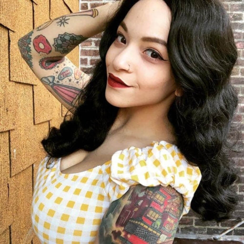 Woman with tattoos wearing a yellow and white checkered top against a wooden and brick wall. Black 1940s style wig with long marcel waves: Vivienne