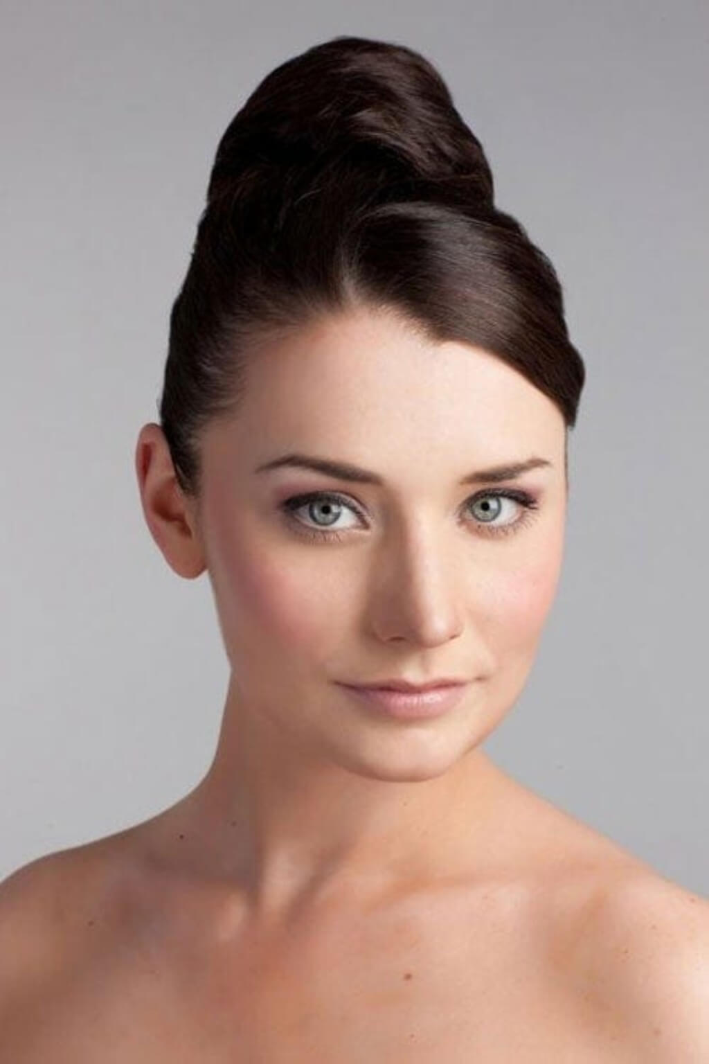 A close-up portrait of a woman with light blue eyes and dark brown hair styled in a high, voluminous bun, set against a solid light grey background. Hairpiece bun; instant perfect bun hairpiece: Hepburn