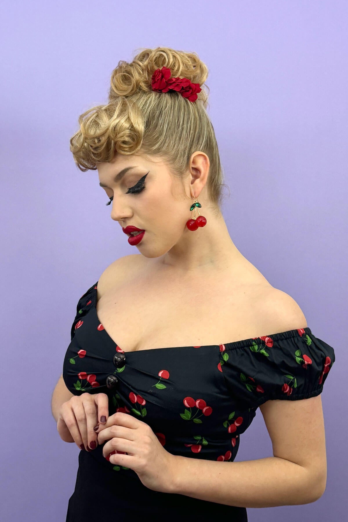 Woman wearing a black dress with red cherry pattern against a purple background. Curly bun hairpiece, rockabilly style: Toni blonde 613T24