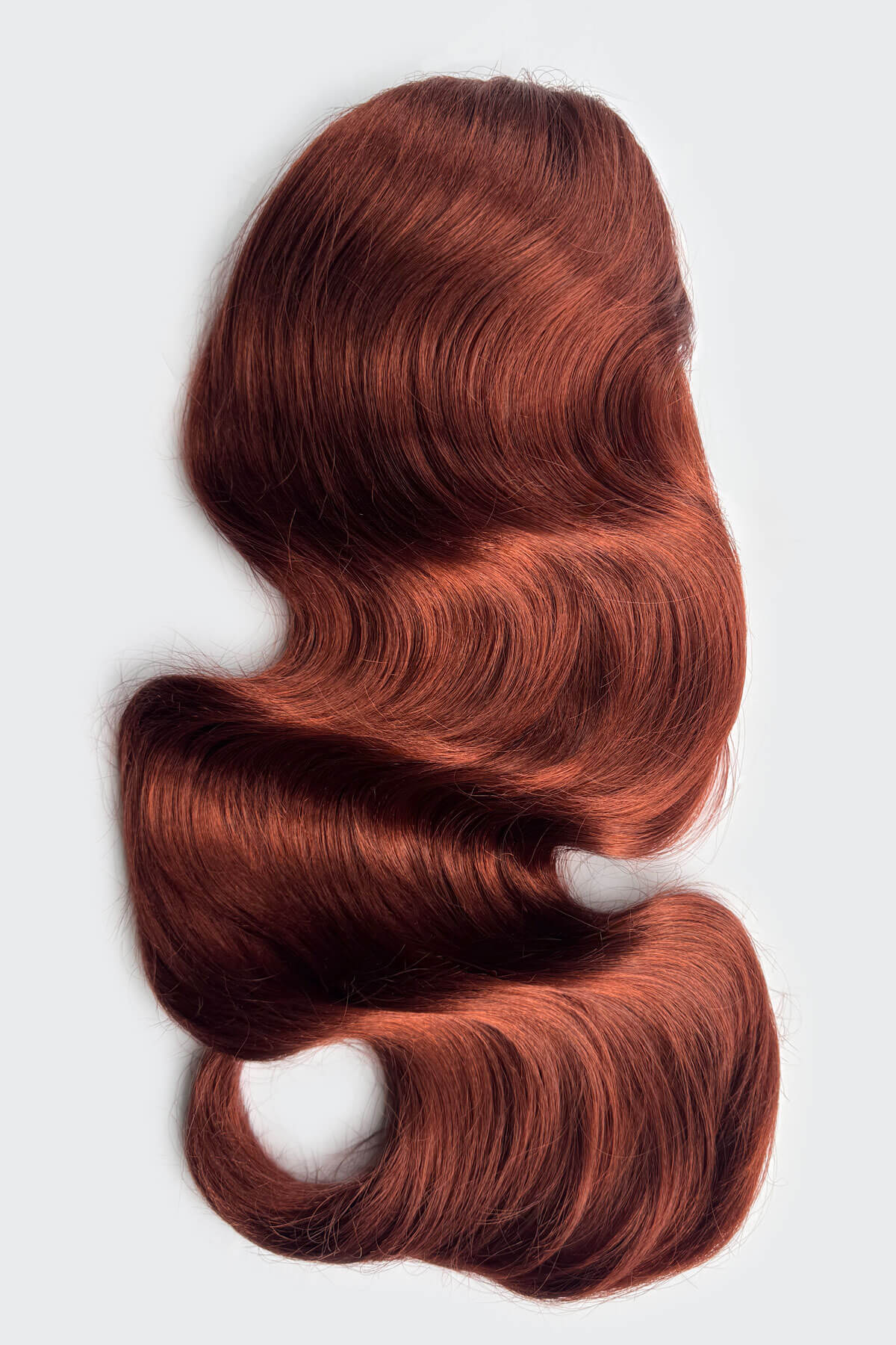 A wavy, hair piece in a warm coppery auburn shade, presented on a light grey background. Copper red hair extension on a white background. Double volume hairpiece extension: Naomi copper red 130