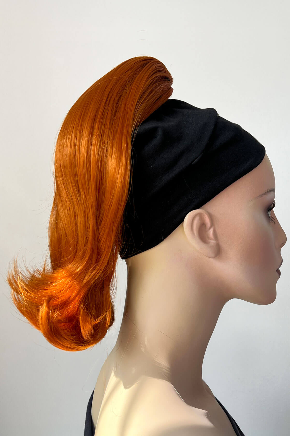 Mannequin head with orange hair styled in a ponytail and black headband against a white background. Ponytail hairpiece, shoulder length with flick/flip, retro 1960s style: Ariana Ginger