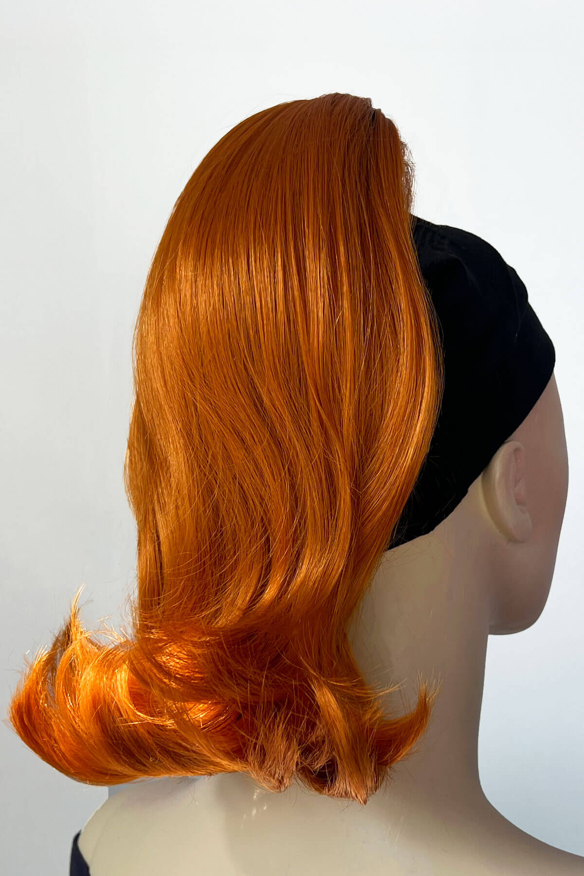 Mannequin head with orange wig and black headband on a white background. Ponytail hairpiece, shoulder length with flick/flip, retro 1960s style: Ariana Ginger