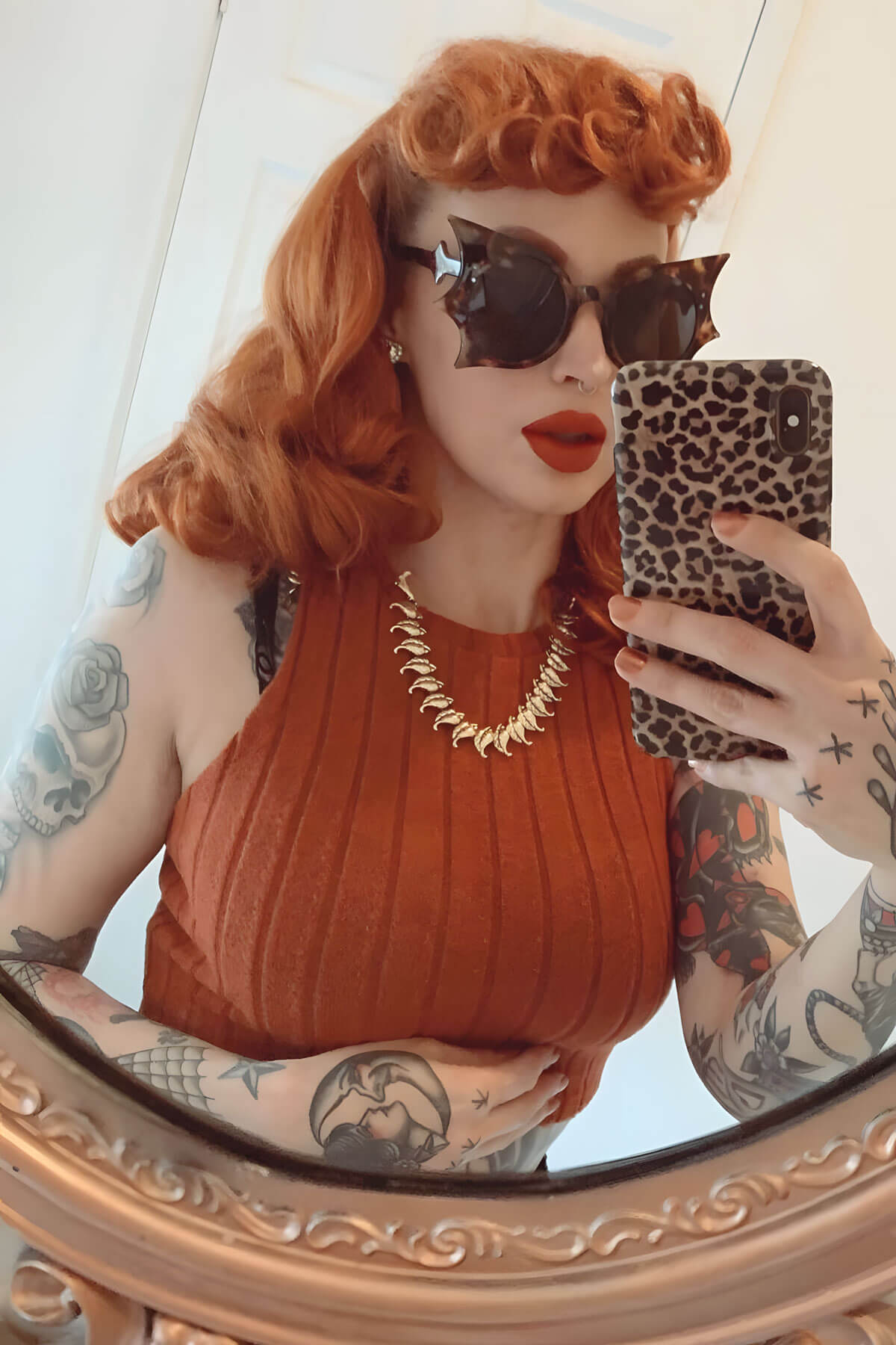 Person with red hair and tattoos taking a mirror selfie wearing sunglasses and an orange top. Curly 1940s style clip-in fringe hairpiece/clip-in bangs: Bunny ginger