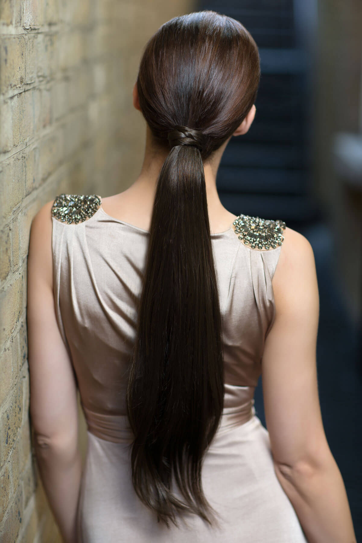 Woman with long dark hair styled in a high ponytail, wearing a light-colored dress with decorative shoulder embellishments. Long sleek ponytail extension with gentle, layered waves: Mali chestnut brown 8