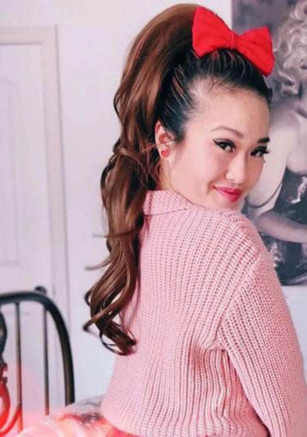 A woman with long brown hair styled in a high ponytail with a large red bow, wearing a pink ribbed knit sweater, photographed indoors. Clip-in ponytail hairpiece extension: Olivia mocha brown 10