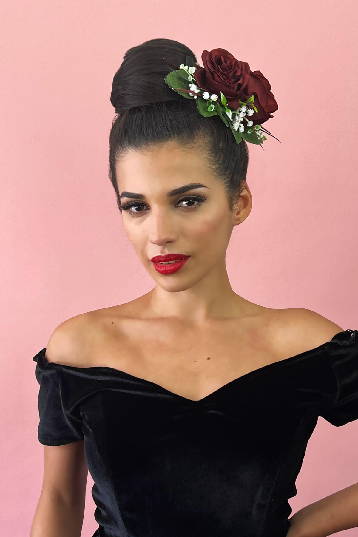 Woman with flowers in her hair wearing a black off-shoulder top against a pink background. Hairpiece bun; instant perfect bun hairpiece: Hepburn