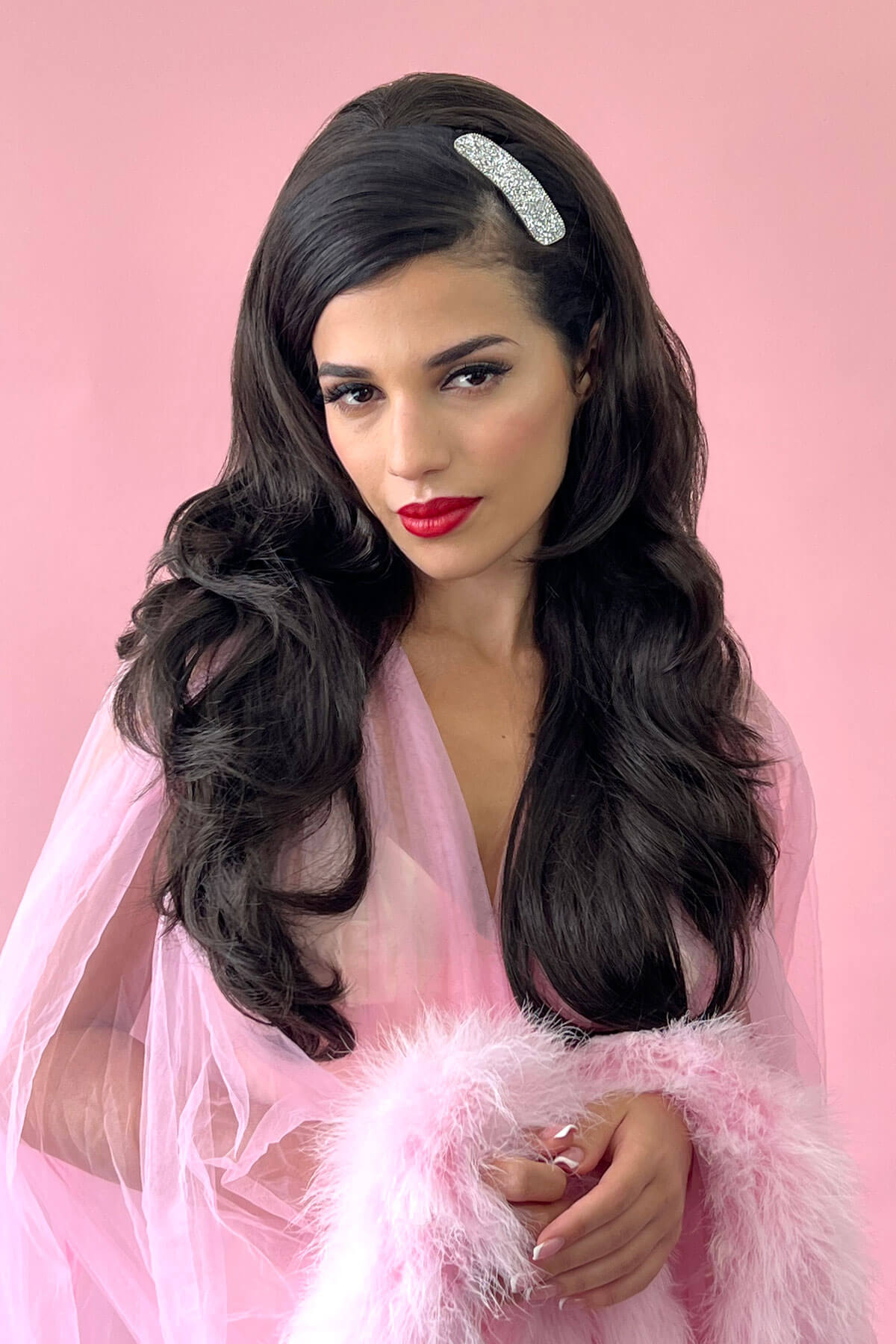 Woman with long dark hair and red lipstick wearing a pink dress against a pink background. 99J