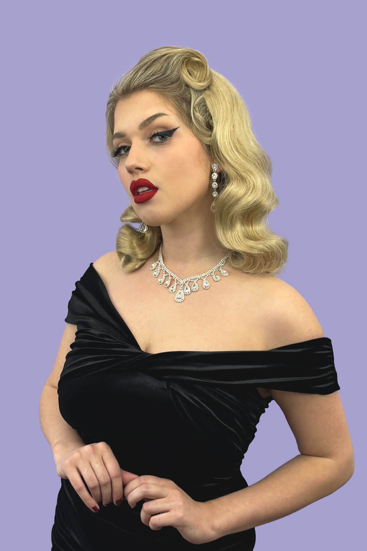 Woman wearing a black off-shoulder dress with a necklace against a purple background. 1950s style half wig hairpiece with deep finger waves: Deborah blonde 613T24