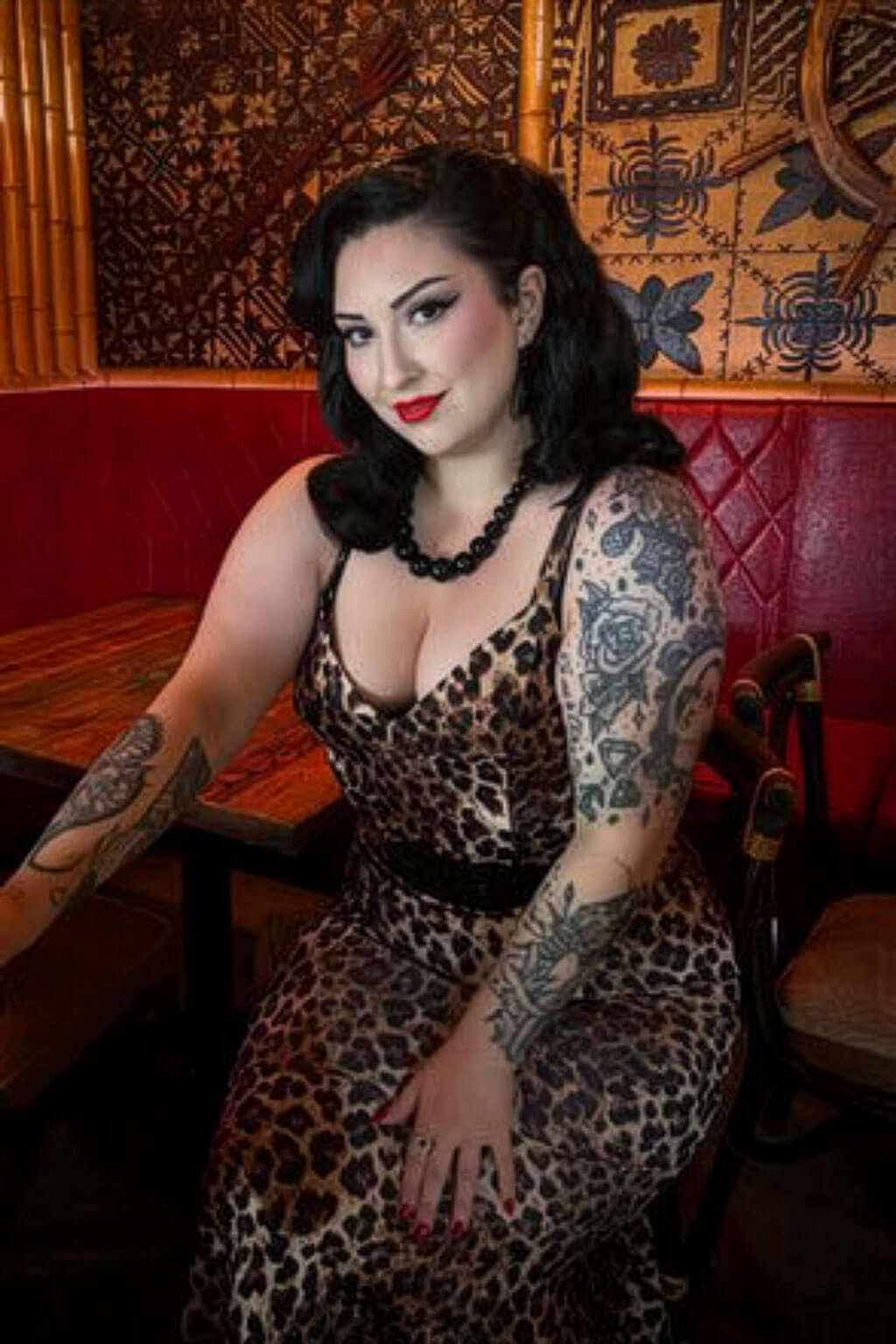 Woman with tattoos wearing a leopard print dress in a decorated room. 1950s style shoulder length half wig hairpiece with beautiful vintage waves: Onyx colour black 1B