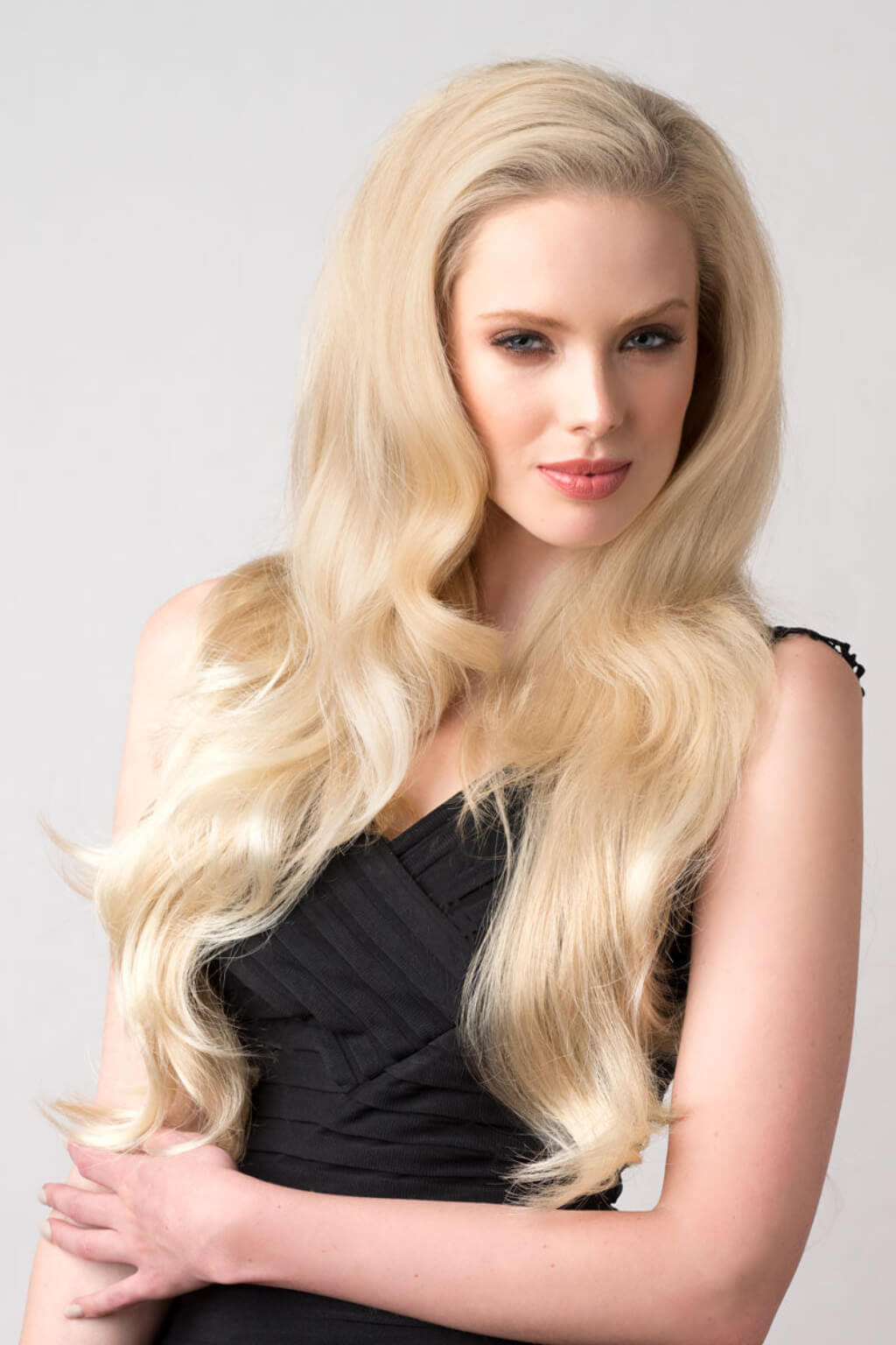 Woman with long blonde hair wearing a black top against a grey background. Blonde half wig hairpiece (3/4 wig), loose curls: Suki blonde 613T24