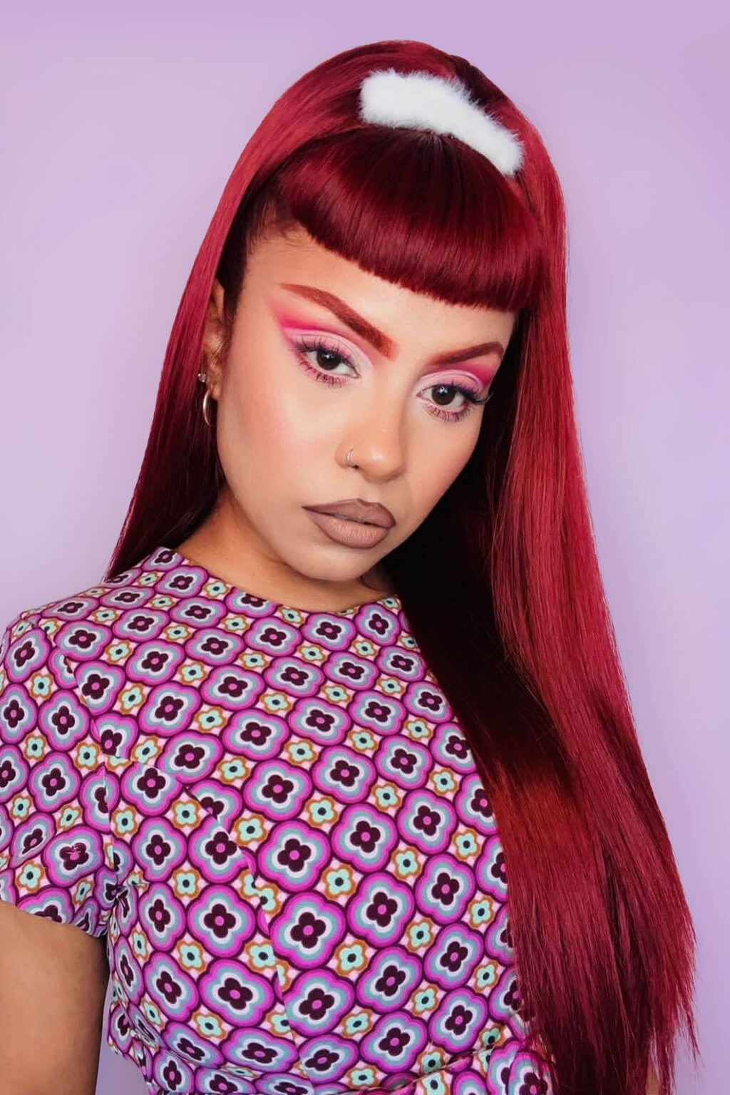 Person with vibrant red hair and colorful makeup wearing a patterned shirt against a light purple background