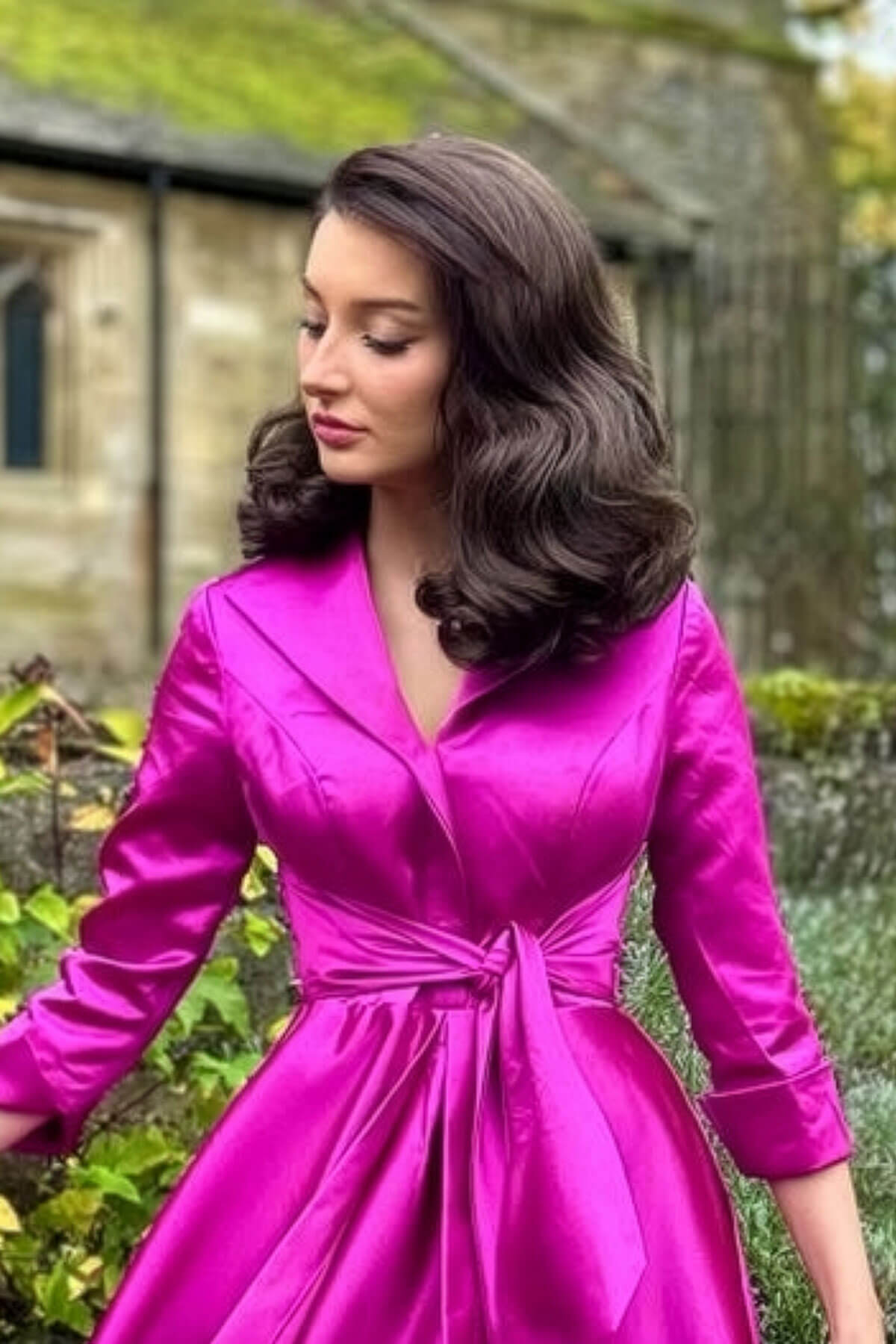 Woman in a bright pink dress standing outdoors with greenery in the background. 1940s style half wig hairpiece with beautiful marcel waves: Virginia chestnut brown 8