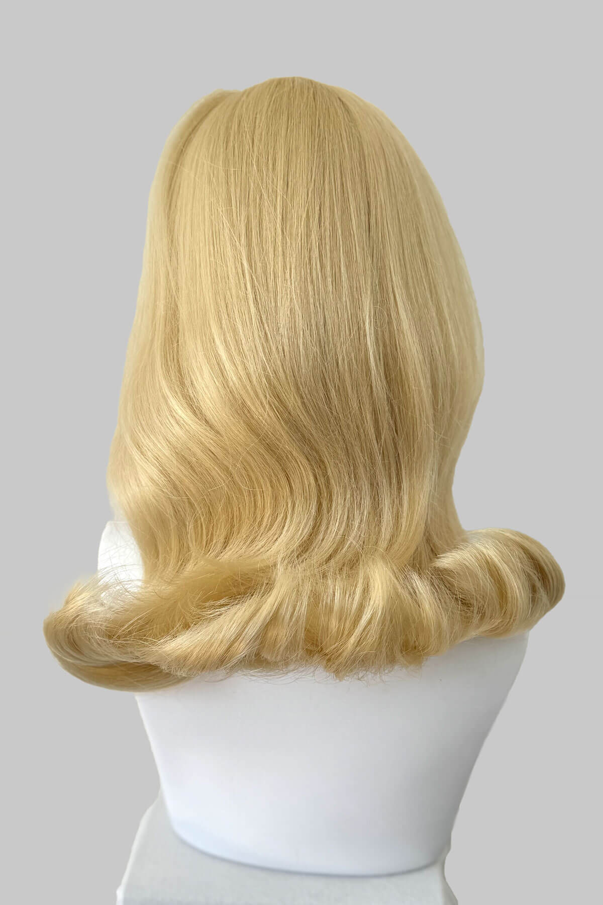 Blonde wig on a mannequin head against a gray background. 1960s half wig hairpiece, flicked, hair flip: Philippa blonde 613