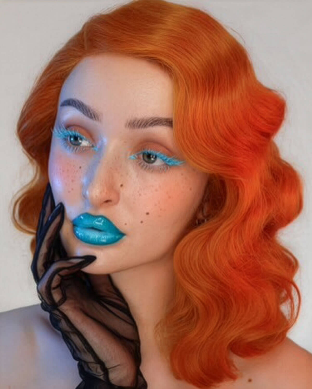 Person with vibrant orange hair and blue makeup on a plain background. Ginger pinup lace front wig: Lula