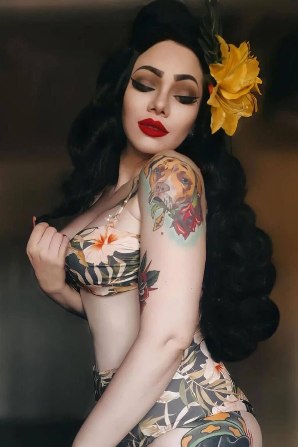 Woman with tattoos and a yellow flower in her hair against a dark background. Long black pinup style wig with vintage hollywood waves: Veronica