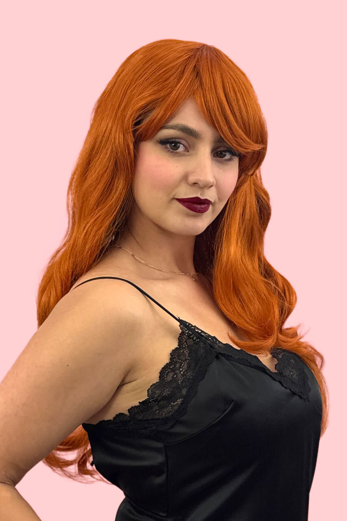 Woman with red hair wearing a black lace top against a pink background. Long ginger wig with loose waves and curls: Ginger