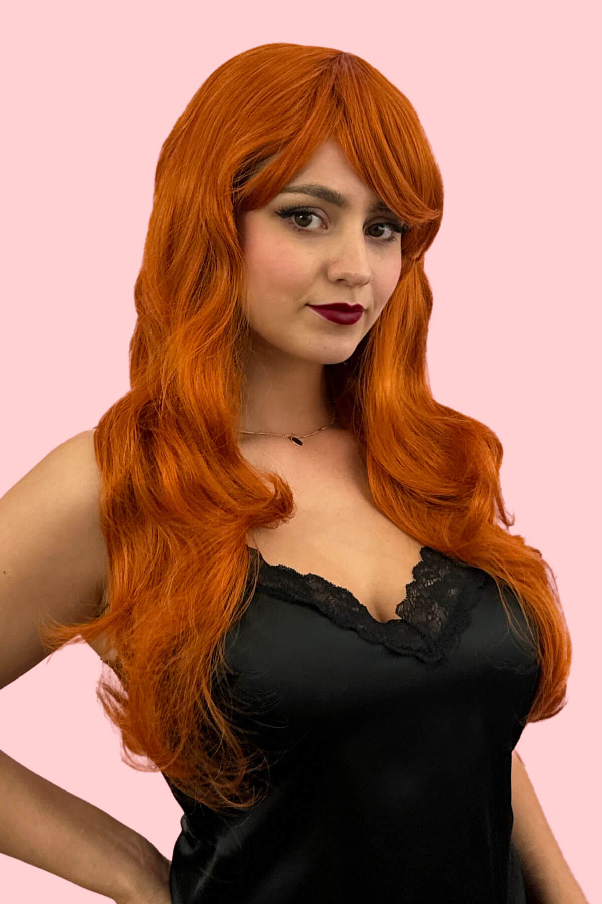 Woman with long orange wig wearing a black top on a pink background. Long ginger wig with loose waves and curls: Ginger