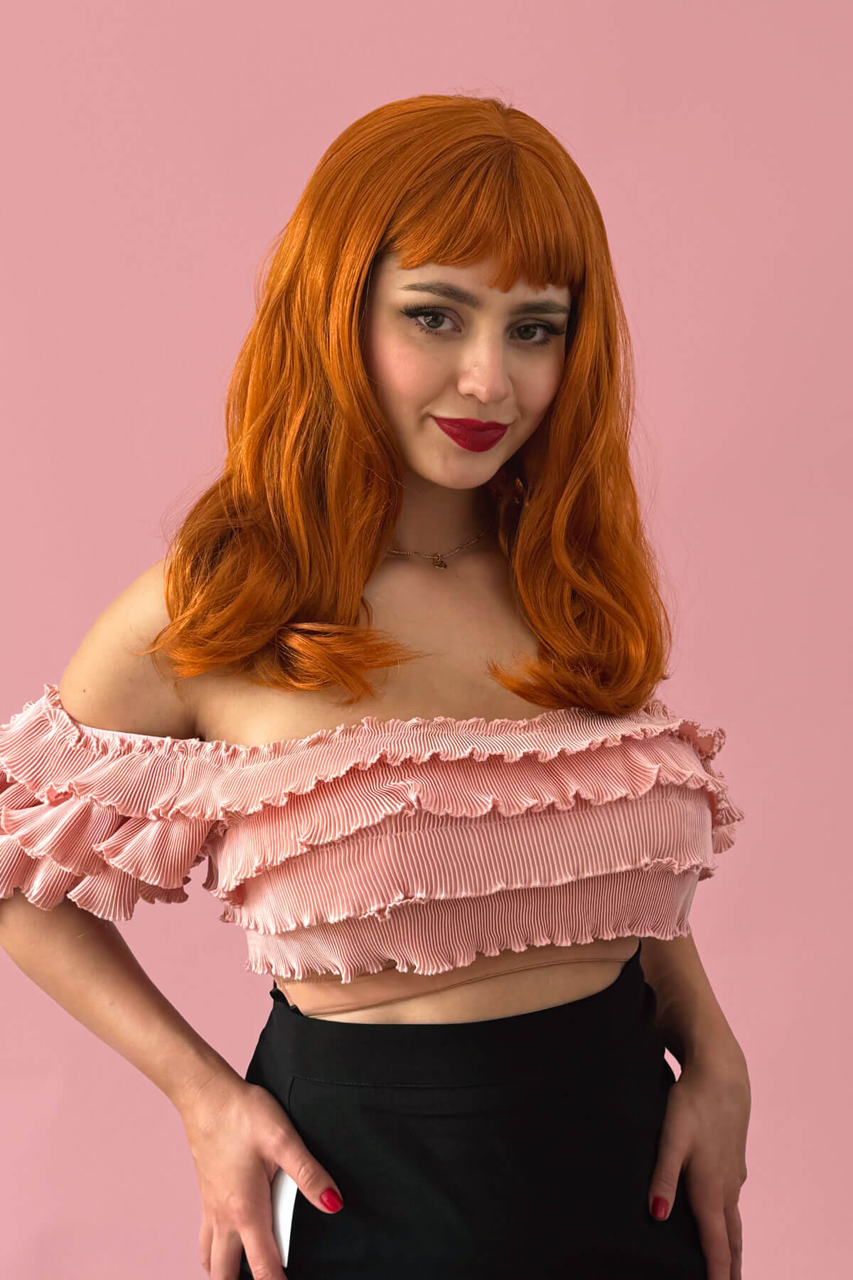Person with ginger hair wearing a pink ruffled top against a pink background. Ginger long wavy bob (LOB) wig with straight retro fringe: Cerissa