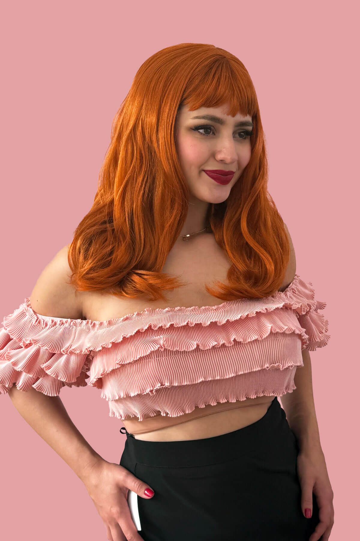 Person with ginger hair wearing a pink ruffled top against a pink background. Ginger long wavy bob (LOB) wig with straight retro fringe: Cerissa