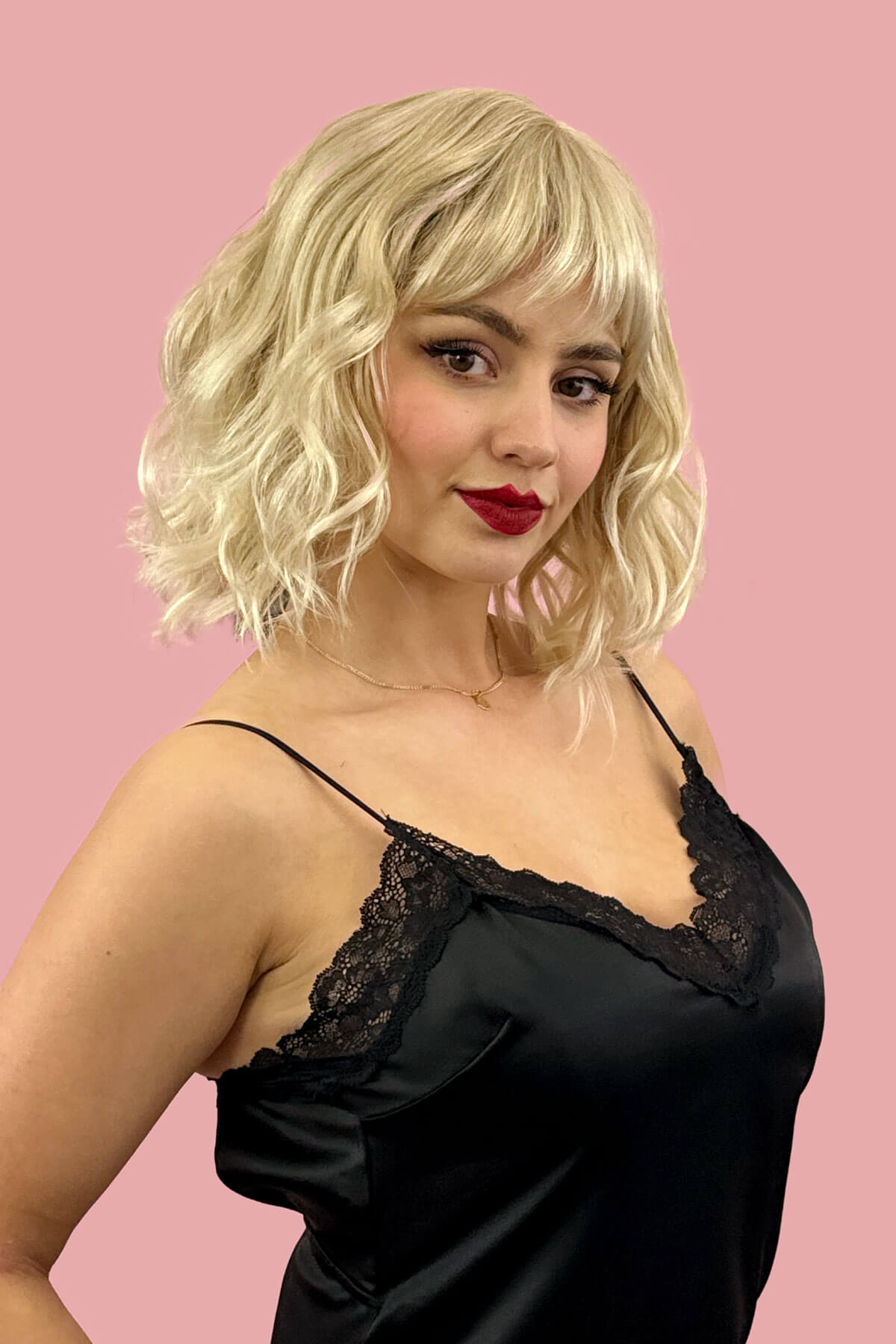 Woman wearing a black lace-trimmed dress against a pink background. Blonde long wavy bob (lob) wig, ash blonde blending to light blonde: Pearl blonde 613T24