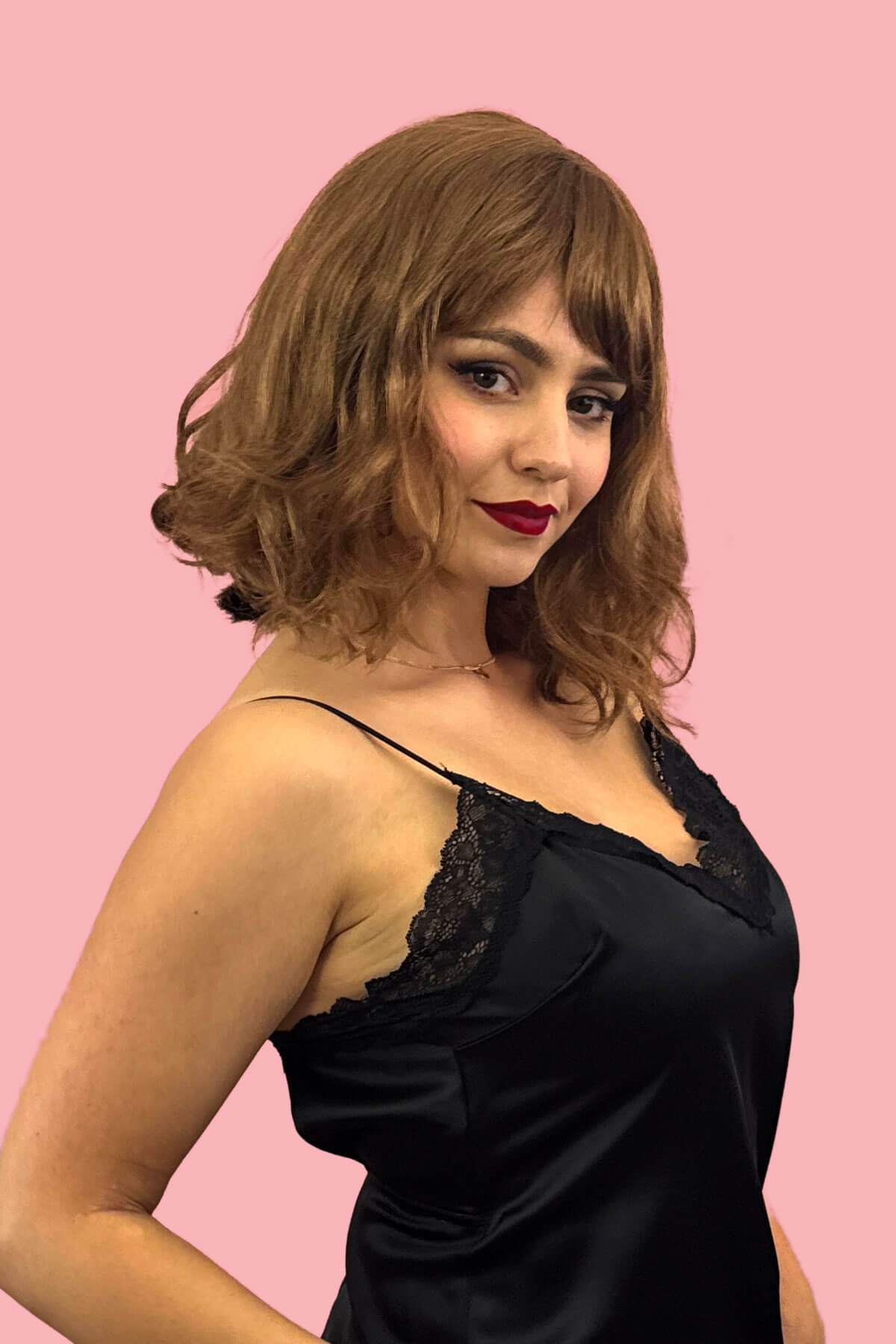 Woman wearing a black satin nightgown with lace details against a pink background. Brown long wavy bob (lob) wig with swept fringe: Clover
