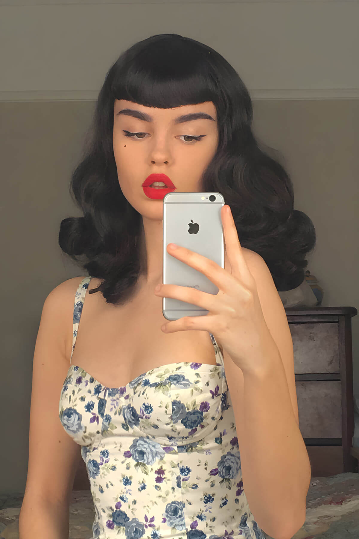 Woman taking a mirror selfie wearing a floral dress and black wig. Black pinup style wig, curled with short fringe, 1950s style: Bettie