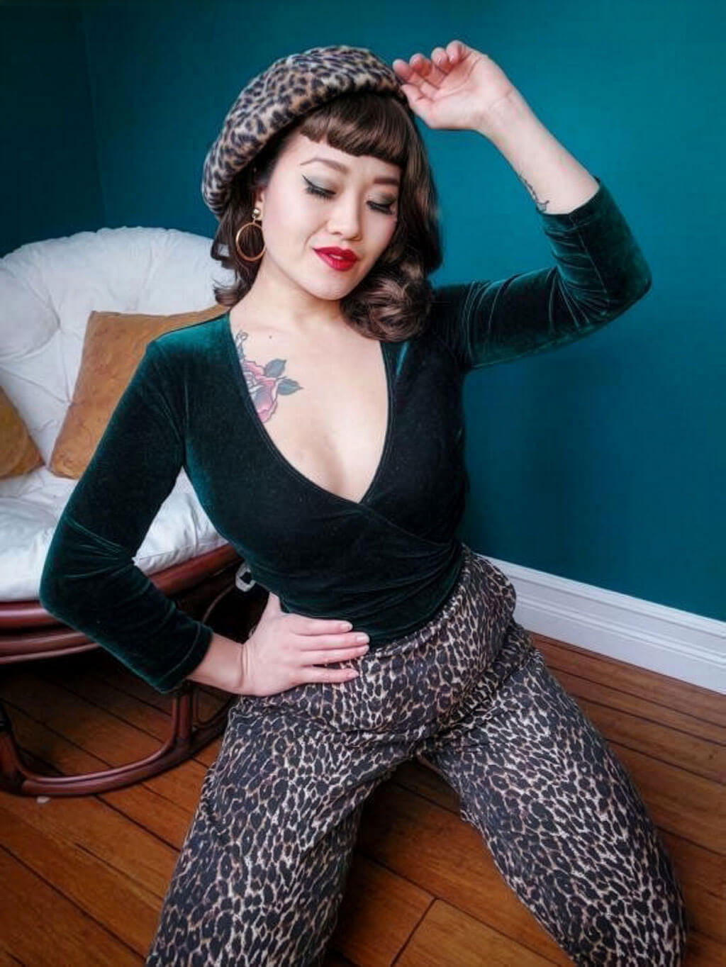 Woman wearing a green velvet top and leopard print pants posing in a room with a blue wall. Medium brown pinup style wig, curled with short fringe: Carolina
