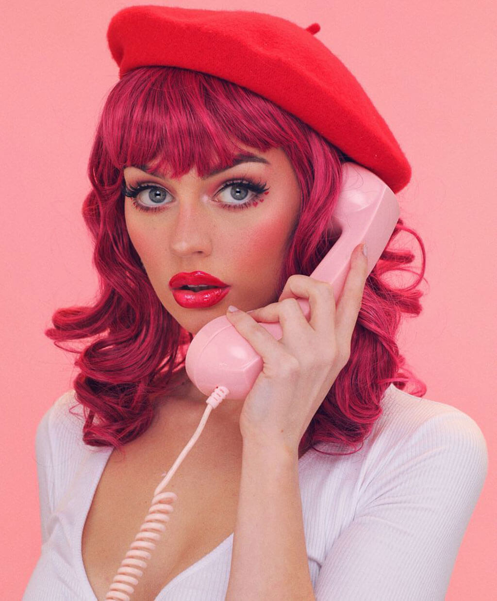 Woman with red hair and a red beret holding a pink phone against a pink background. Black and pink pinup wig, curled with a short fringe, 1950s style: Billie