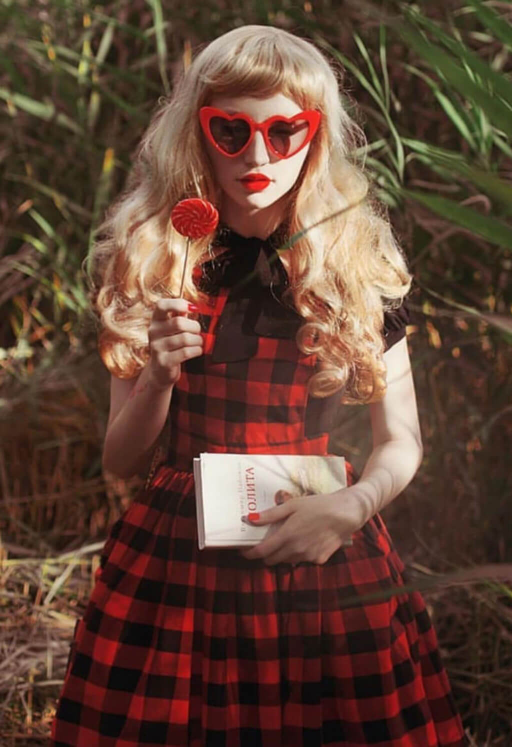 Woman in a red and black checkered dress holding a heart-shaped object and a book. Long blonde 1950s style wig, gently wavy with short fringe: Emmeline