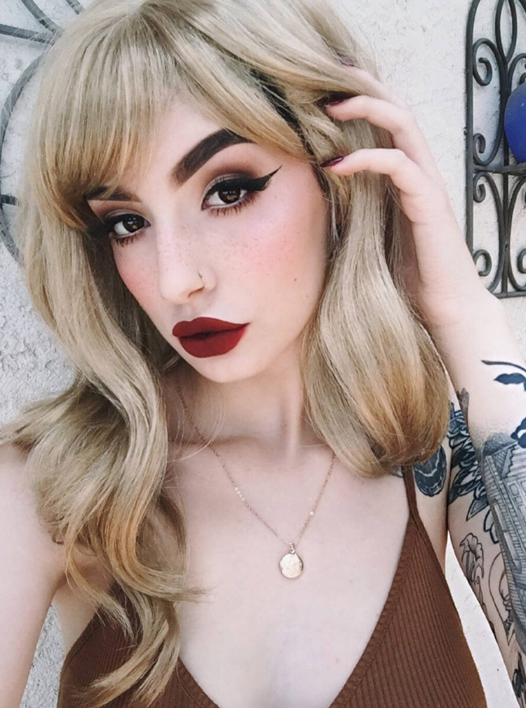 Woman with blonde hair and red lipstick posing against a decorative wall. Long ash blonde wig with gentle waves: Harriet