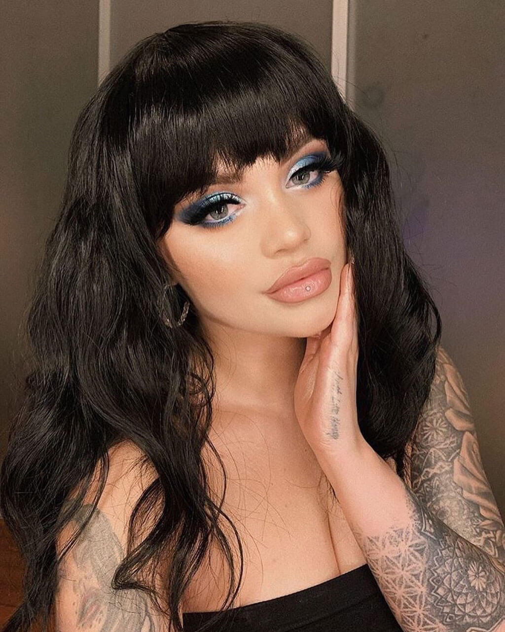Woman with tattoos and black hair posing against a neutral background. Black long wavy bob wig (lob): Sigrid