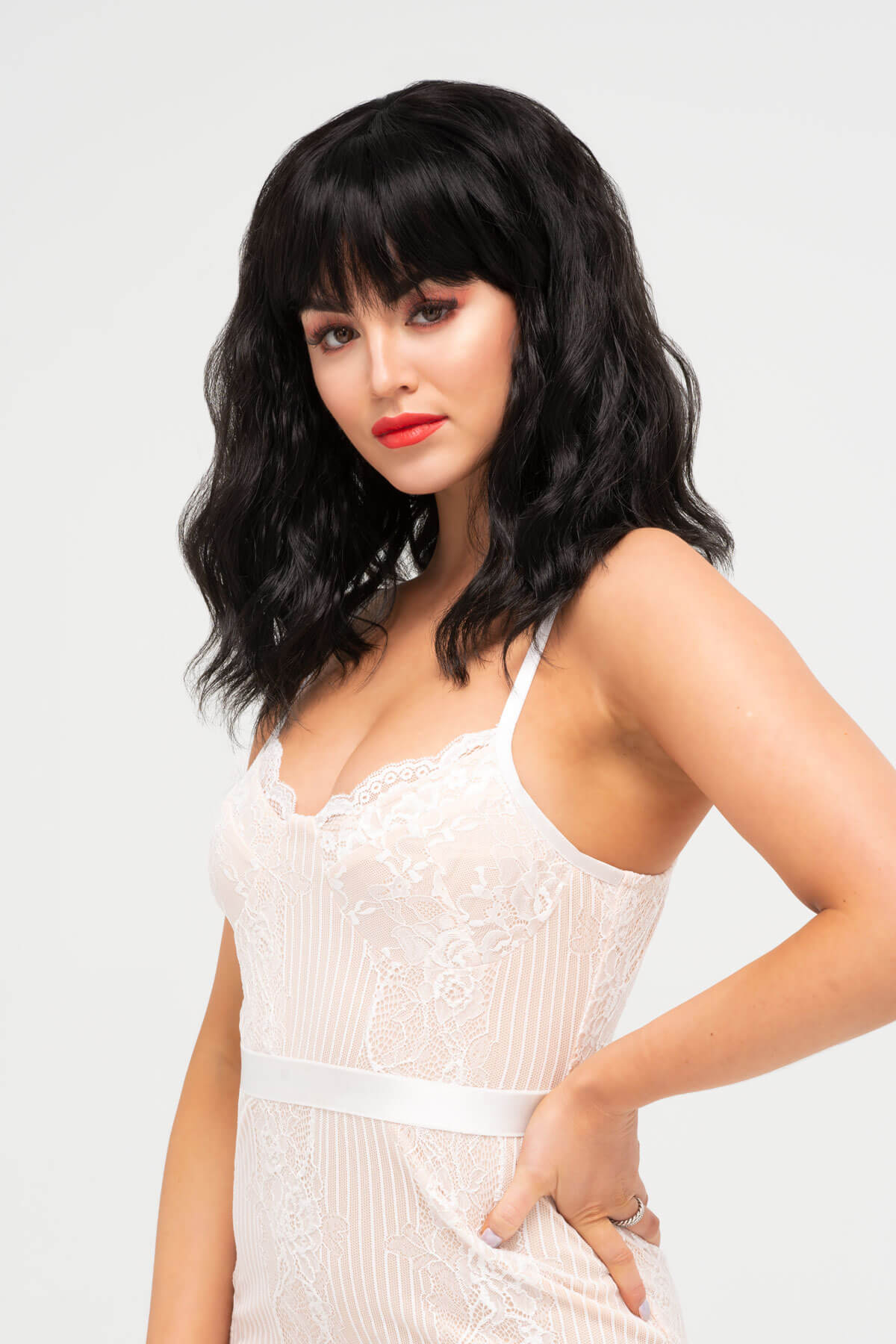 Woman with black hair and red lipstick wearing a white dress against a light gray background. Black long wavy bob wig (lob): Sigrid