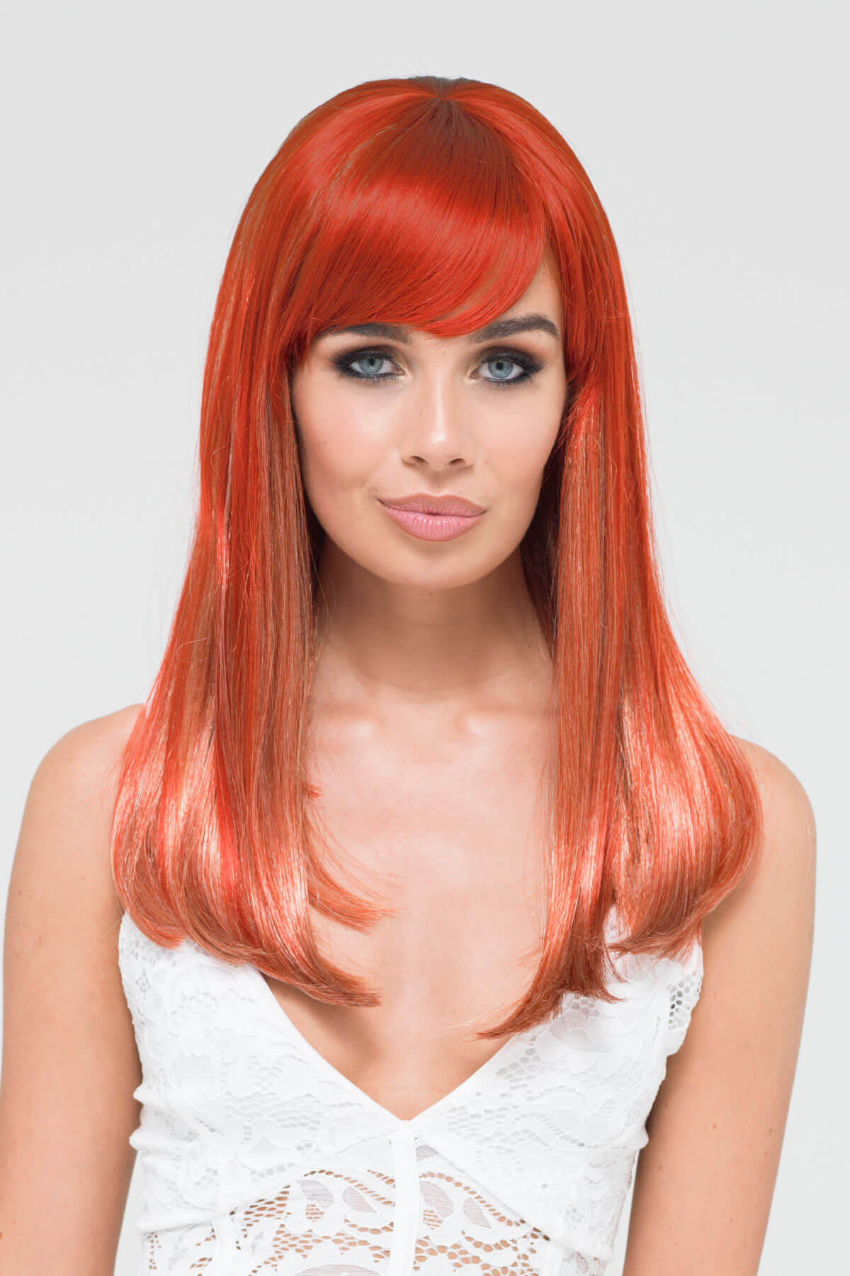 Woman with vibrant red hair and a white lace top on a light gray background. Copper red and blonde wig with straight fringe: Mathilda