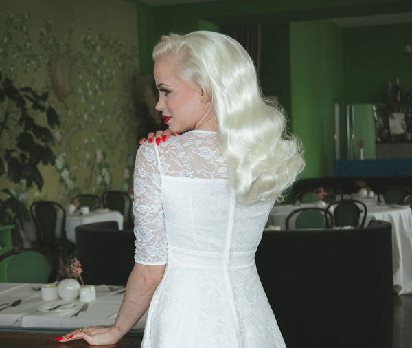 Woman in a white lace wedding  dress with long blonde hair in a room with tables and chairs.
