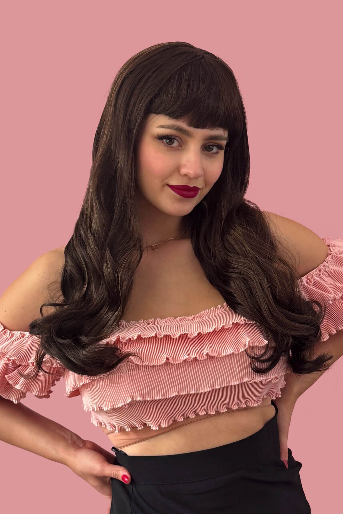 A woman wearing a long chestnut brown wig with blonde highlights and a pinup-style fringe, posing in front of a pink background. Long chestnut brown wig with blonde highlights and pinup-style fringe: Zola