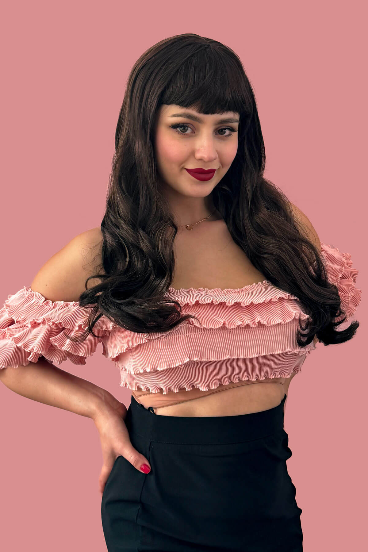 A woman wearing a long chestnut brown wig with blonde highlights and a pinup-style fringe, posing in front of a pink background. Long chestnut brown wig with blonde highlights and pinup-style fringe: Zola