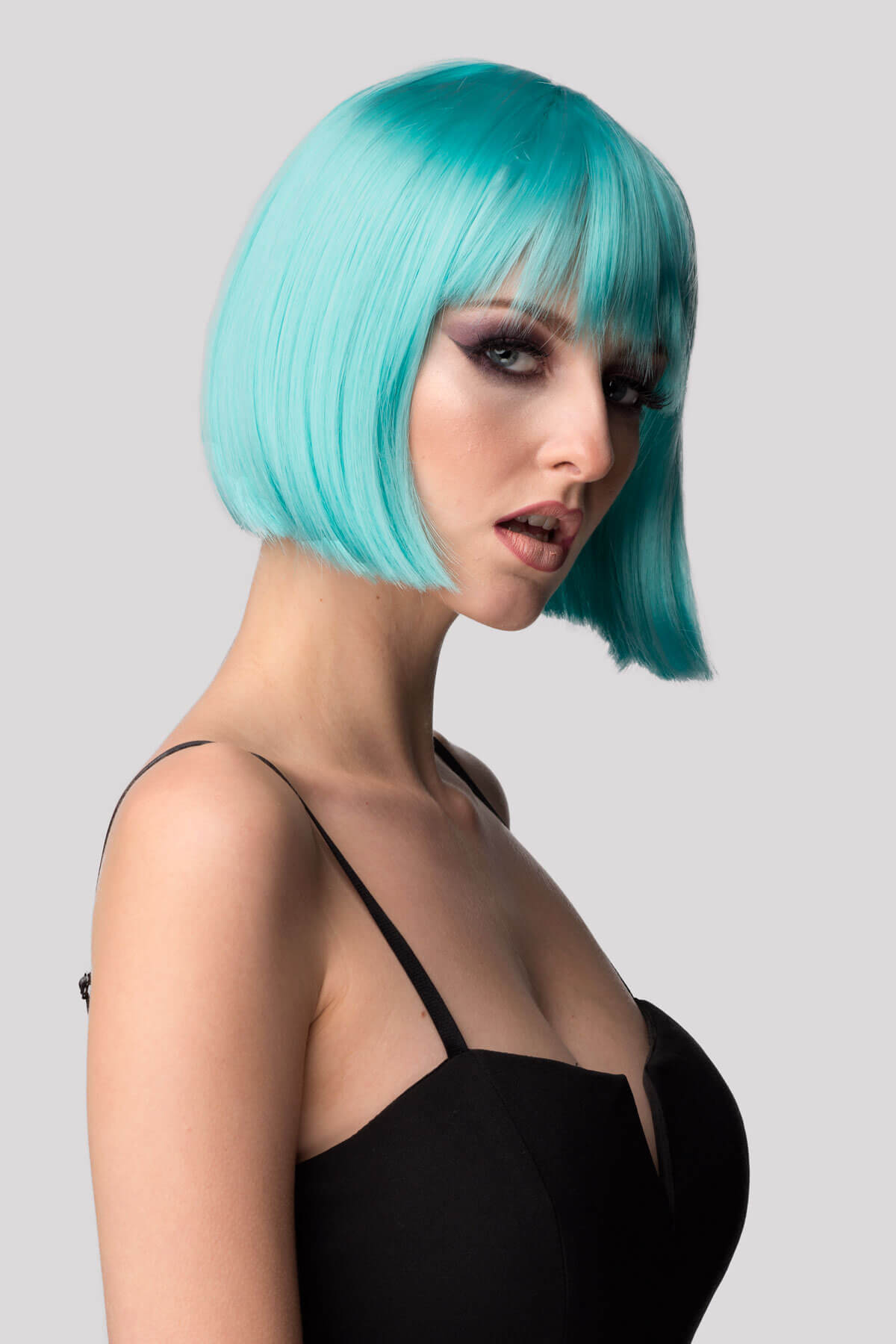 Woman with turquoise hair wearing a black top against a gray background. Turquoise bob wig  with full fringe: Iris