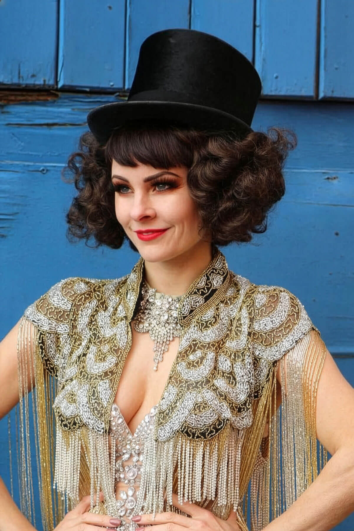 Woman in a vintage outfit with a black hat and beaded top against a blue wooden background. A short brown vintage style wig with waves: Rita brown 8
